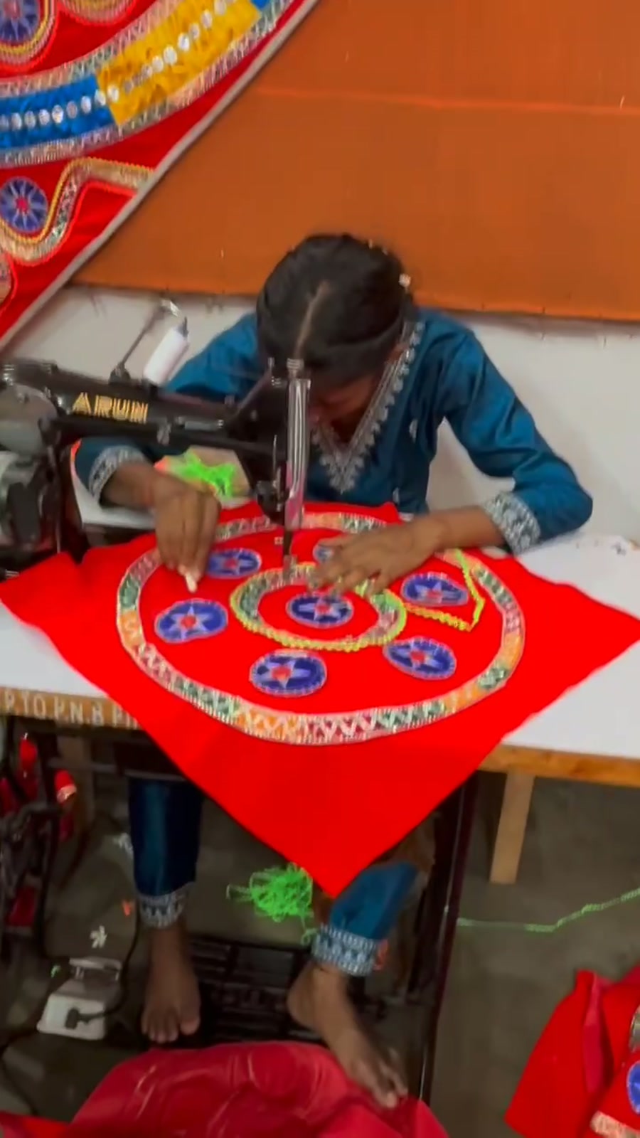 An artisan at a treadle sewing machine stitching mirrored appliqué medallions onto red cloth.