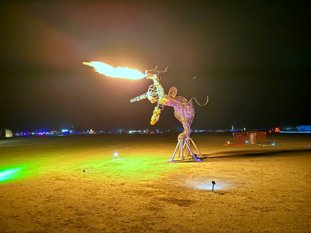 Navagunjara Reborn on the Black Rock Desert playa at night, 2026 — the dragon's head breathing a long jet of flame above the woven body on its tripod base, lit green and gold from the ground.