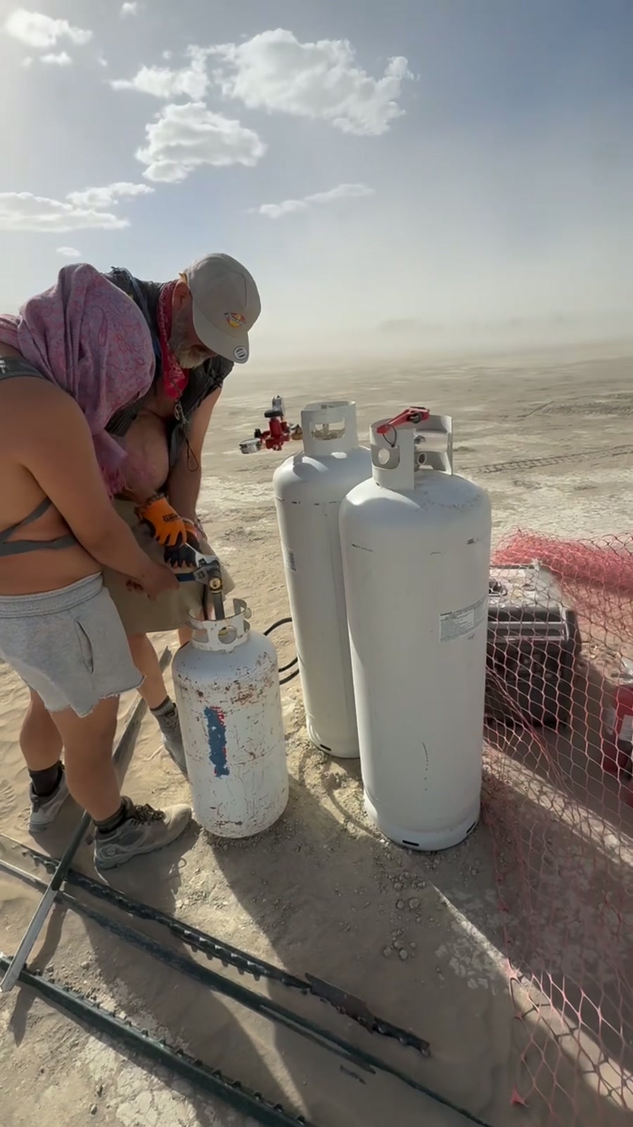 Two crew members connect propane cylinders in blowing dust on the open playa.