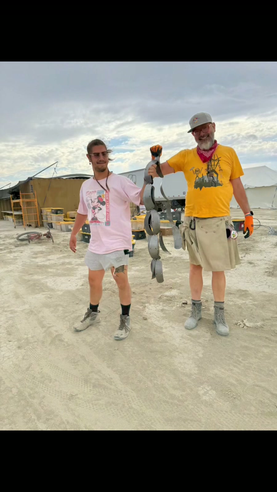 Two crew members hold up a linked chain of cut aluminum scales outside a workshop.