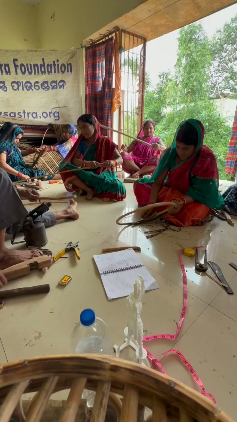 Women in saris seated on a tile floor weaving bamboo cane under a Kalasastra Foundation banner.