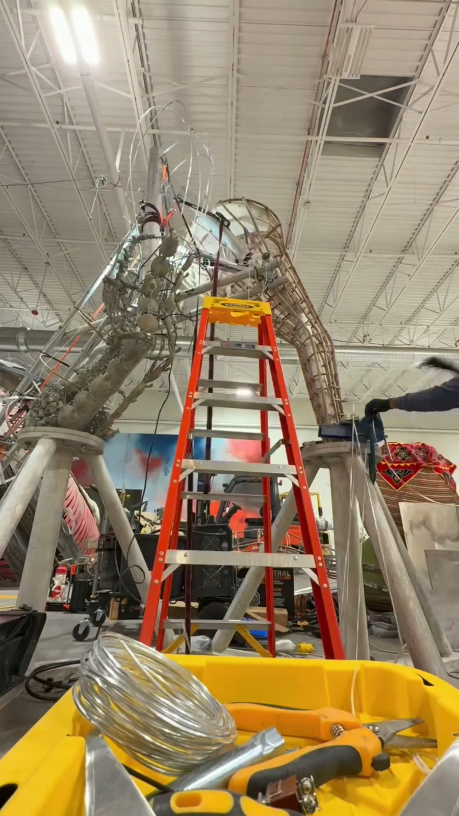 Aluminum truss frame rising inside a workshop beside an orange ladder; wire and tin snips in a yellow bin in the foreground.